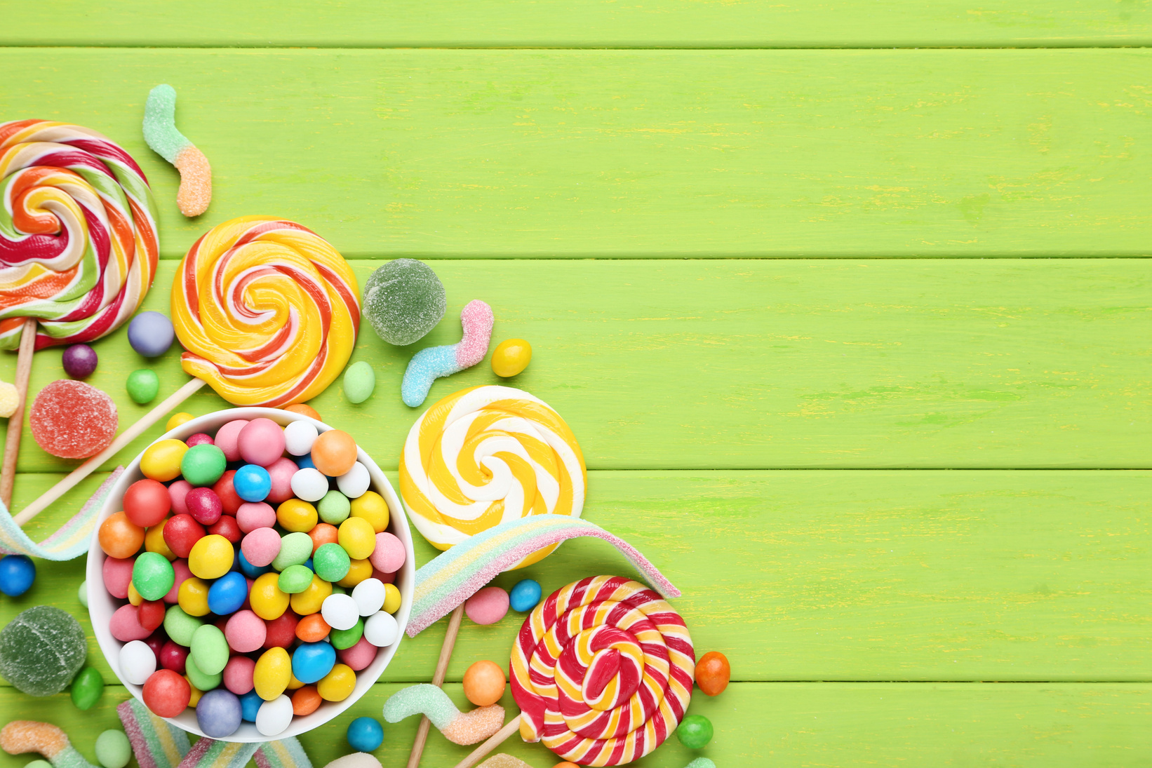 Sweet candies in bowl and lollipops on green wooden table